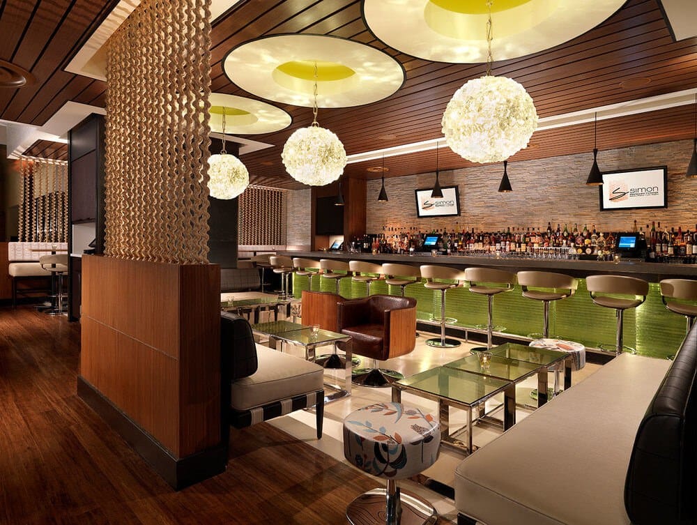 Stylish bar lounge with a long green-tiled bar, cream stools, glass coffee tables, white seating, and oversized hanging spherical lights under a wood-paneled ceiling.