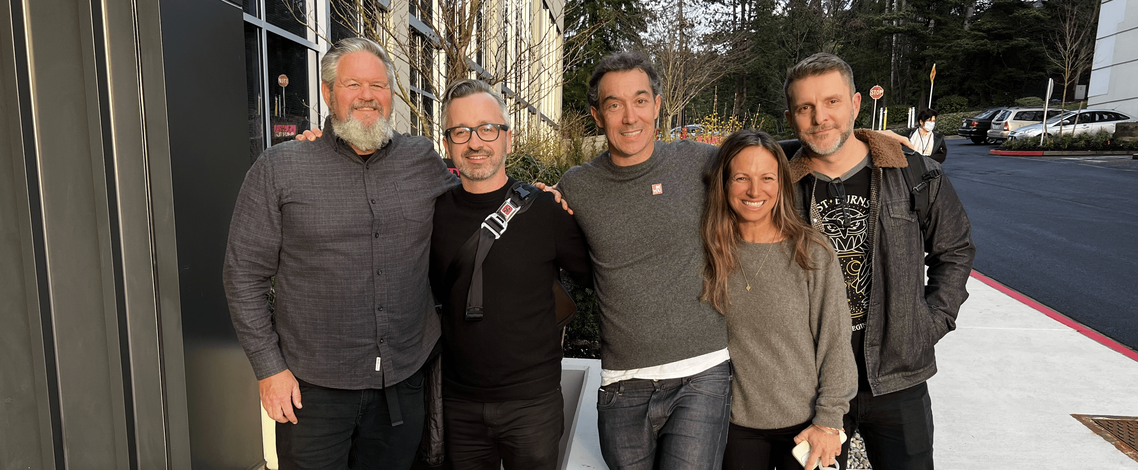 Five people stand together outdoors in front of a building, smiling; wearing casual sweaters and jackets, with trees and parked cars in the background.