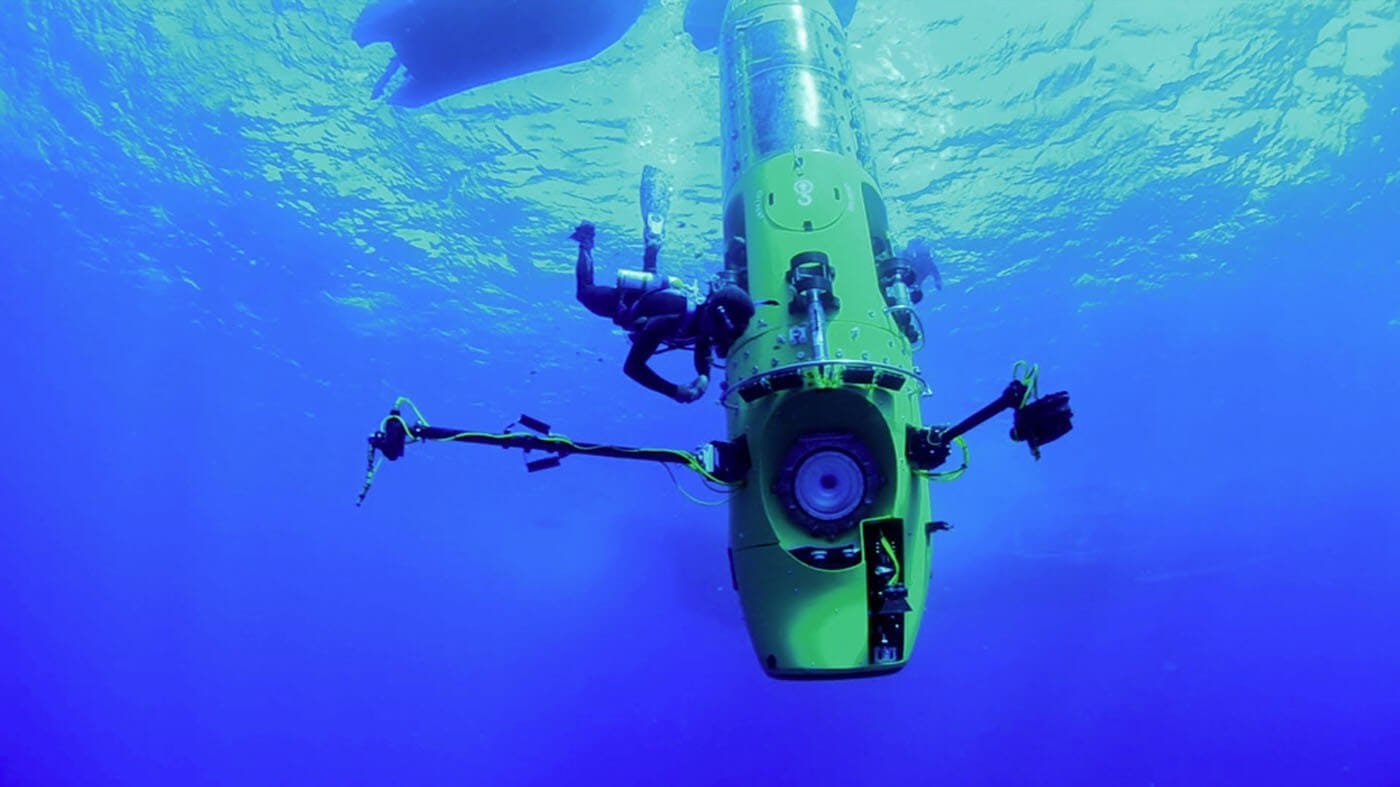 Green underwater remotely operated vehicle with robotic arms in clear blue ocean, surrounded by two divers.