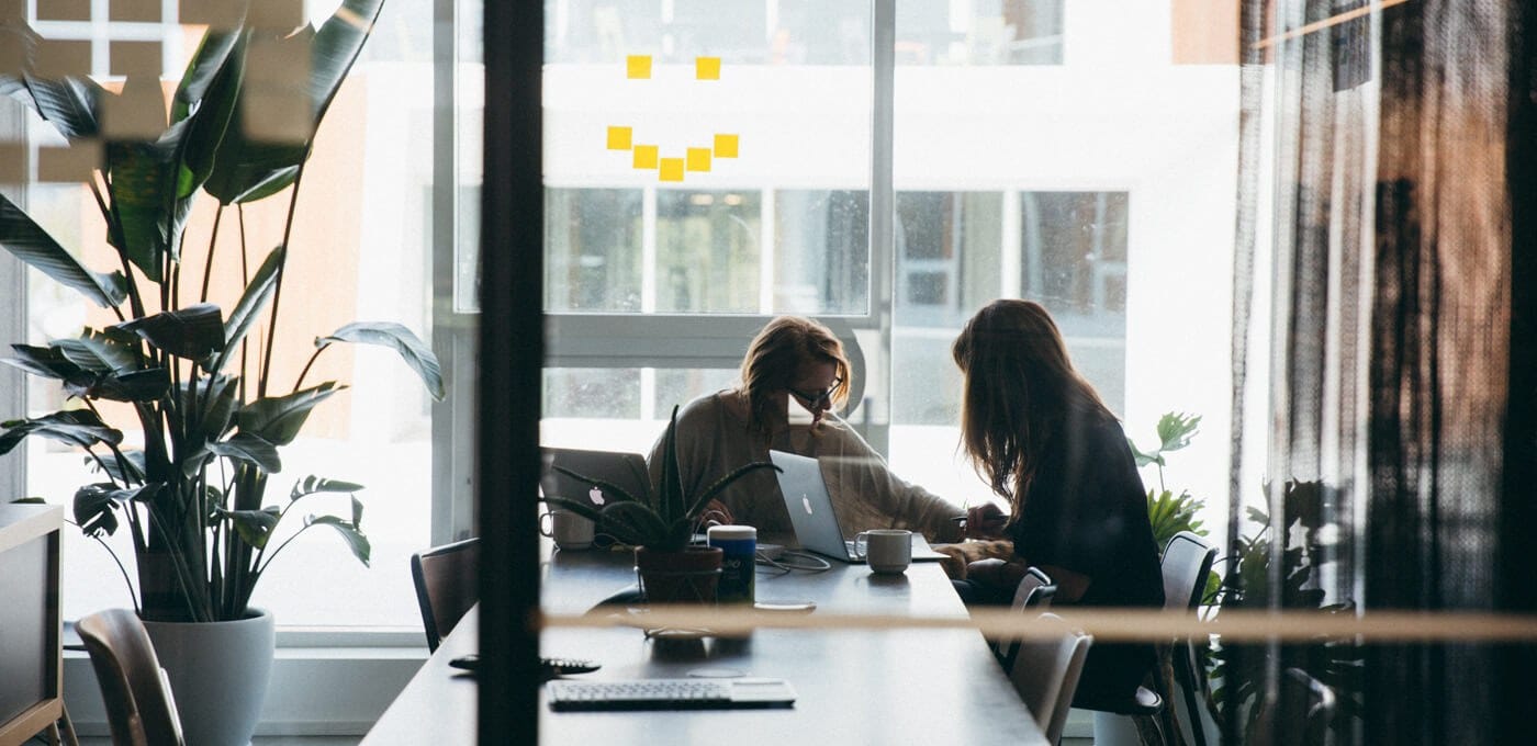 Two people work at a long table with laptops and coffee, surrounded by plants; bright windows and a yellow sticky-note smile on the back wall.