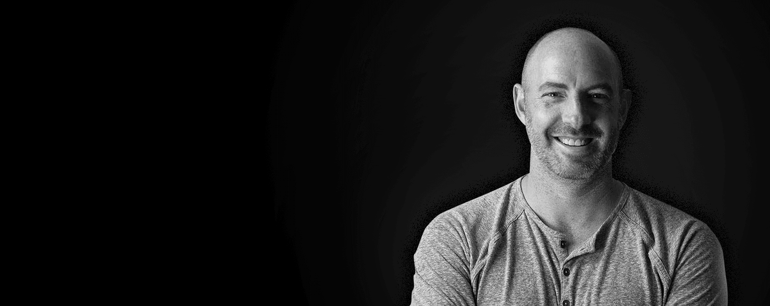 Smiling, bald person with light stubble wearing a gray Henley shirt, against a dark gradient background.