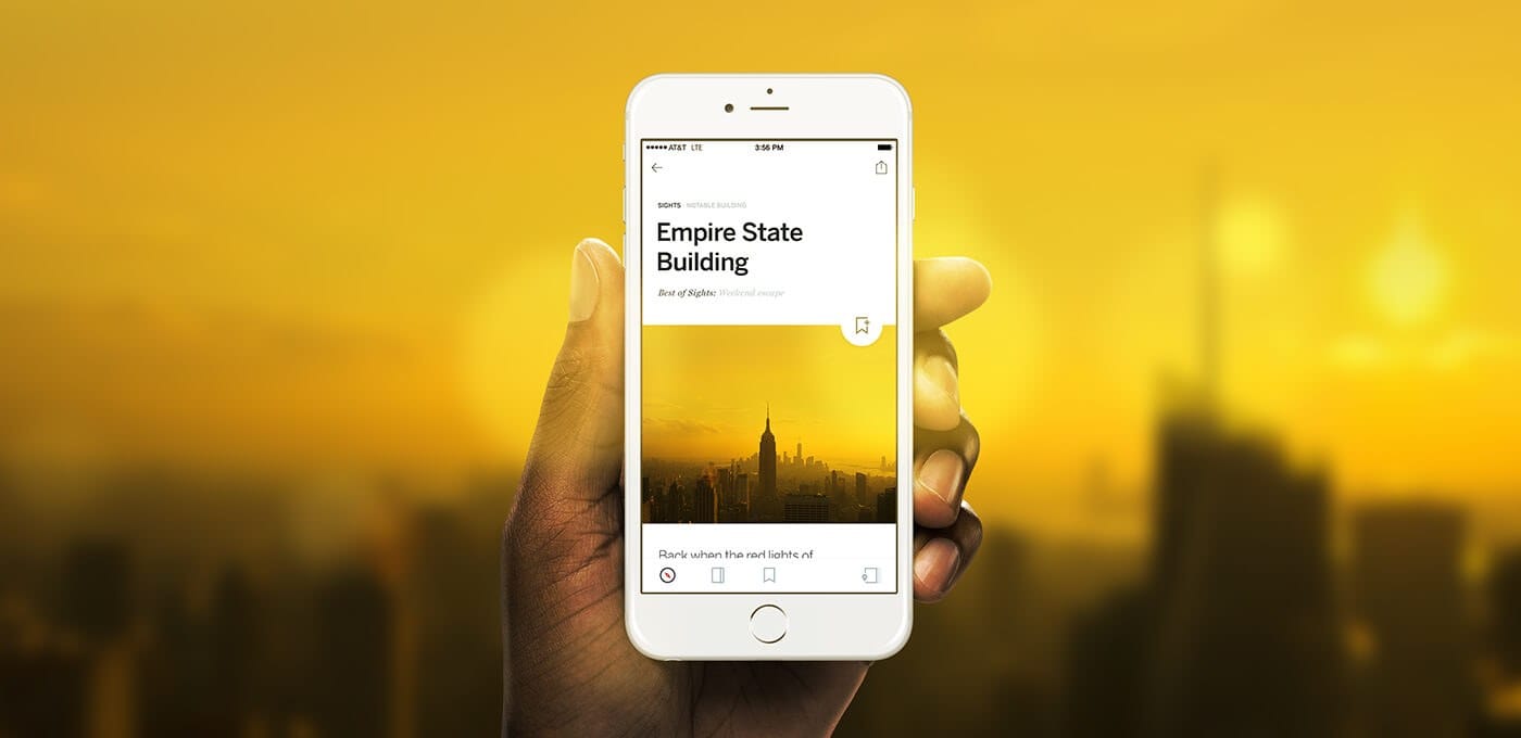 A hand holding a white smartphone showing an article titled “Empire State Building” with a city skyline image, against a warm yellow sunset background.
