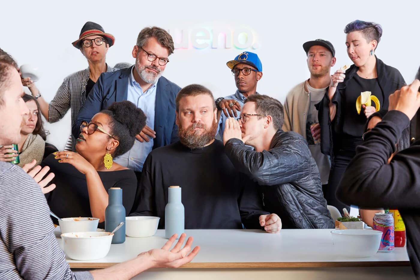 A diverse group around a table with bowls and drinks at a casual gathering; one person whispers to another as others chat.