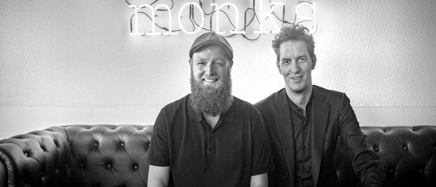 Two adults sit on a leather couch, smiling at the camera; one with a beard and cap, the other in a blazer, beneath a neon 'monks' sign.