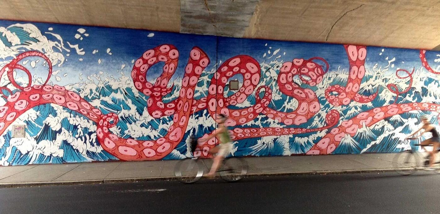 Vibrant mural of pink octopus tentacles curling across blue waves on a wall, with two blurred cyclists passing by in front.