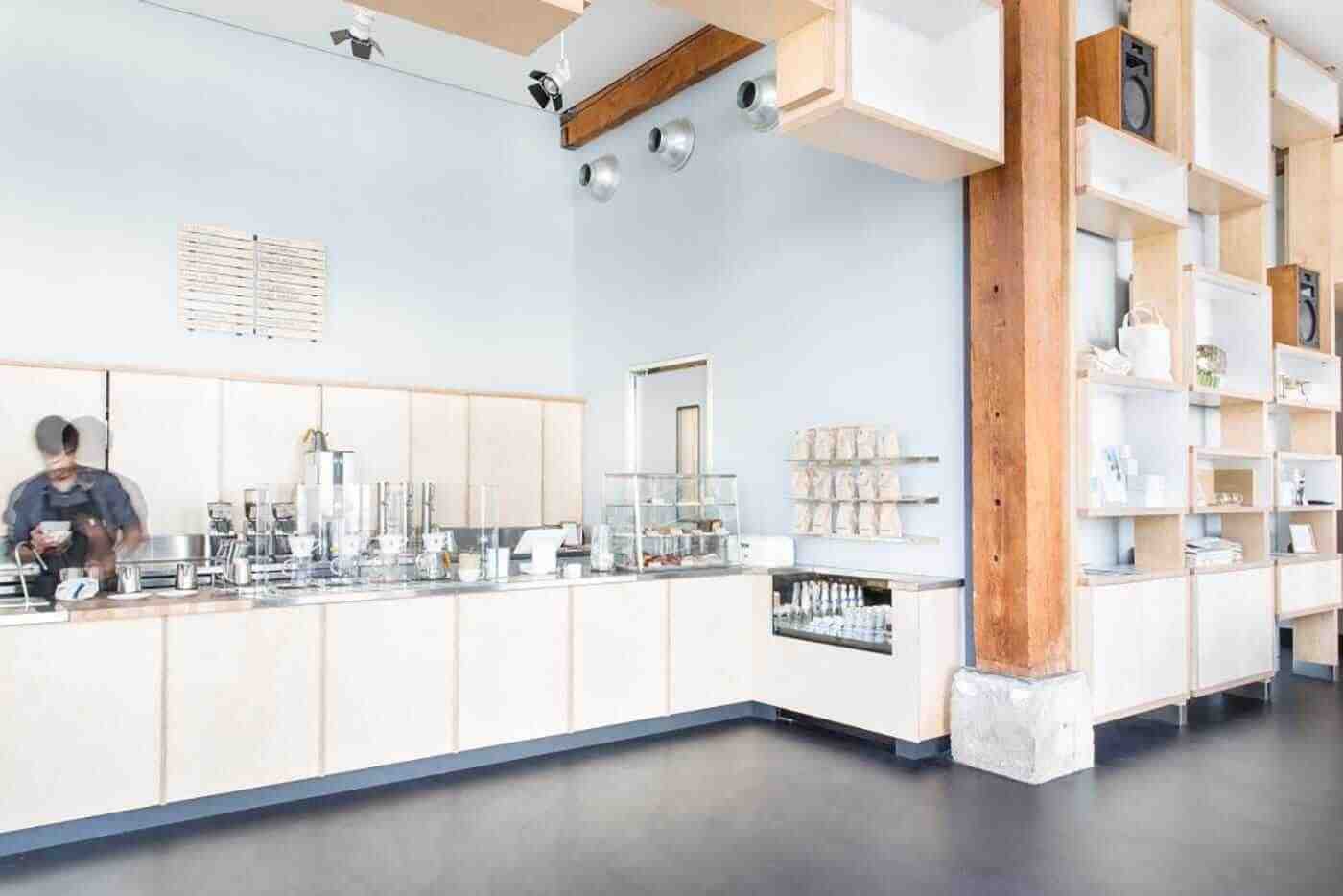 A bright, minimalist cafe with a long pale-wood counter, a barista at work, a glass pastry display, and shelves along the right wall beside a wooden column.