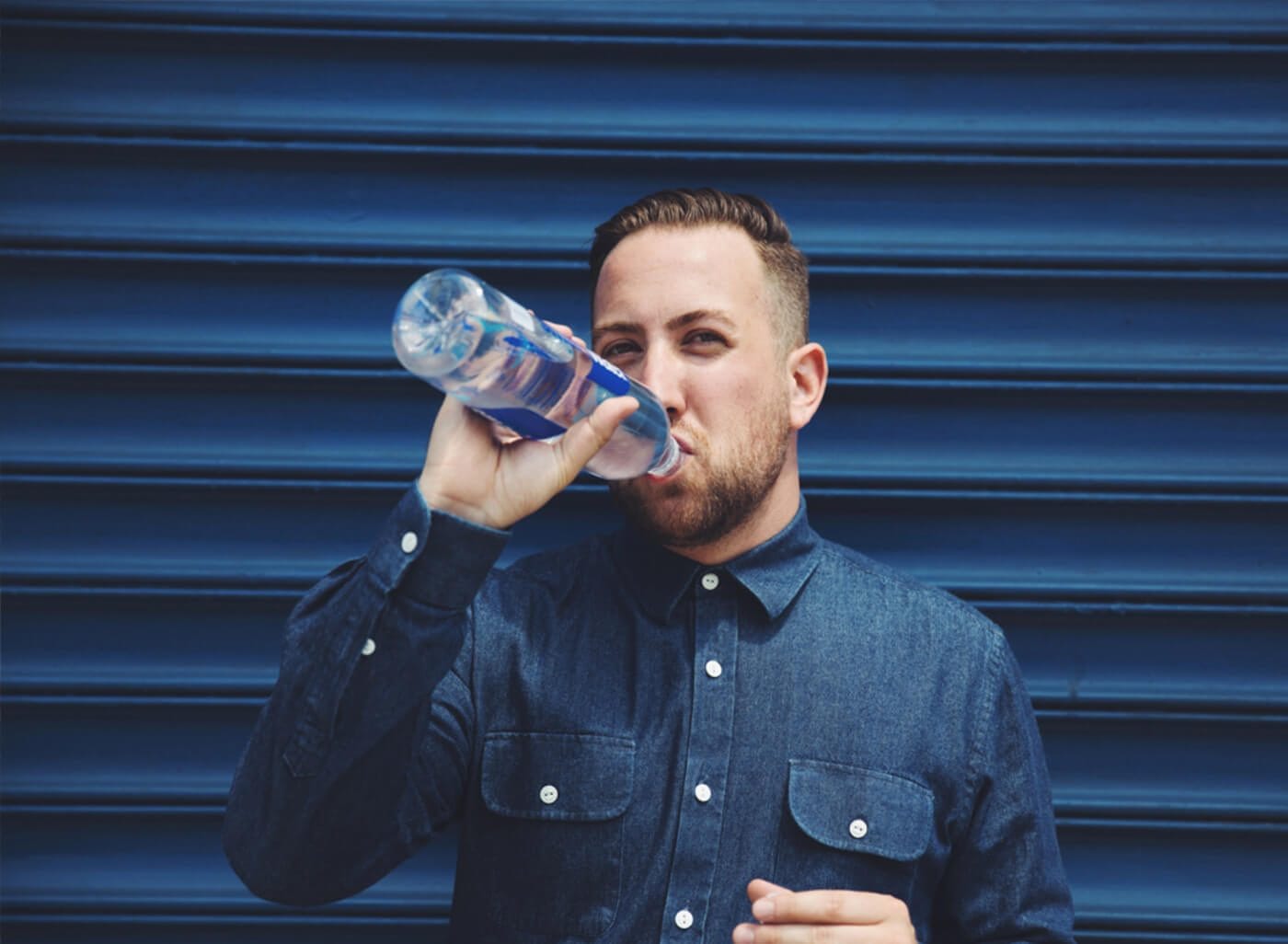 A person with short hair and a beard in a denim shirt drinks from a clear plastic water bottle against a blue corrugated wall.