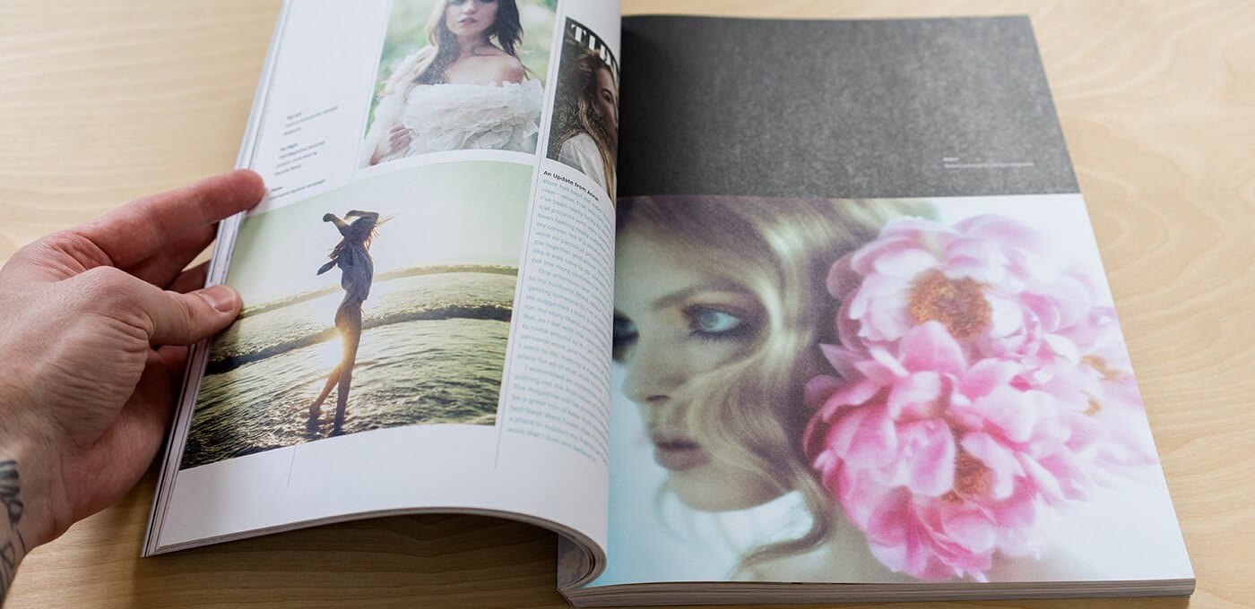 Open magazine on a light wooden table; a tattooed hand turns the pages. Left page shows a person in a white dress by the water, right page a close-up with pink flowers.