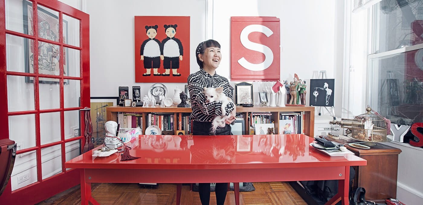Person behind a glossy red desk in a bright, art-filled room, holding a small white fluffy dog, with bold red artwork and shelves in the background.